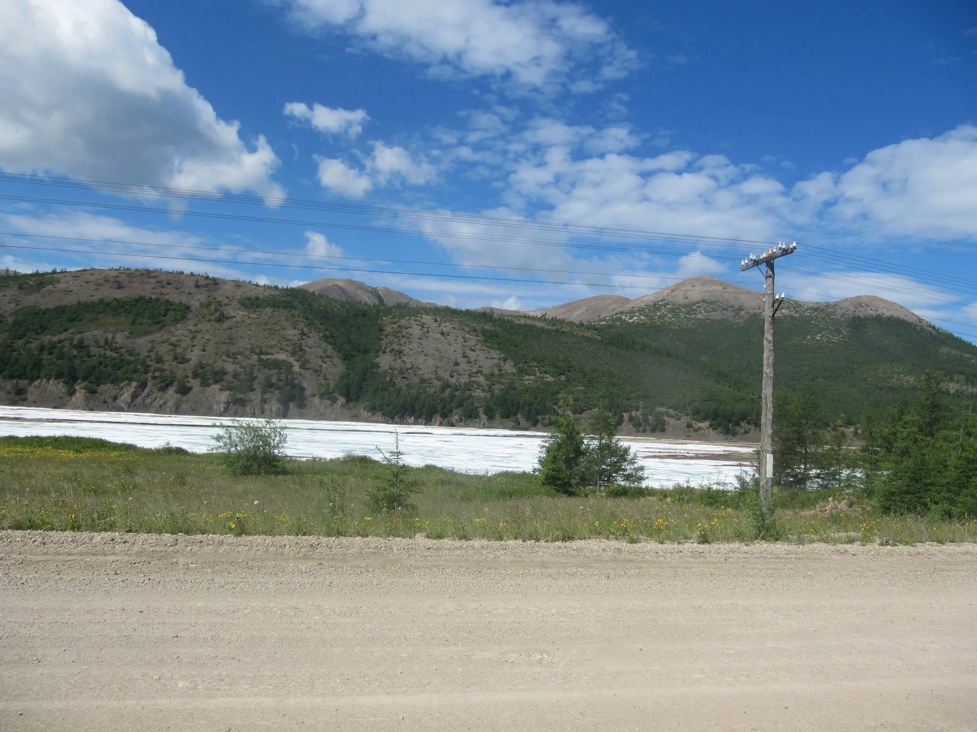 A wide gravel stretch of the Kolyma Highway beside a river with mountains behind