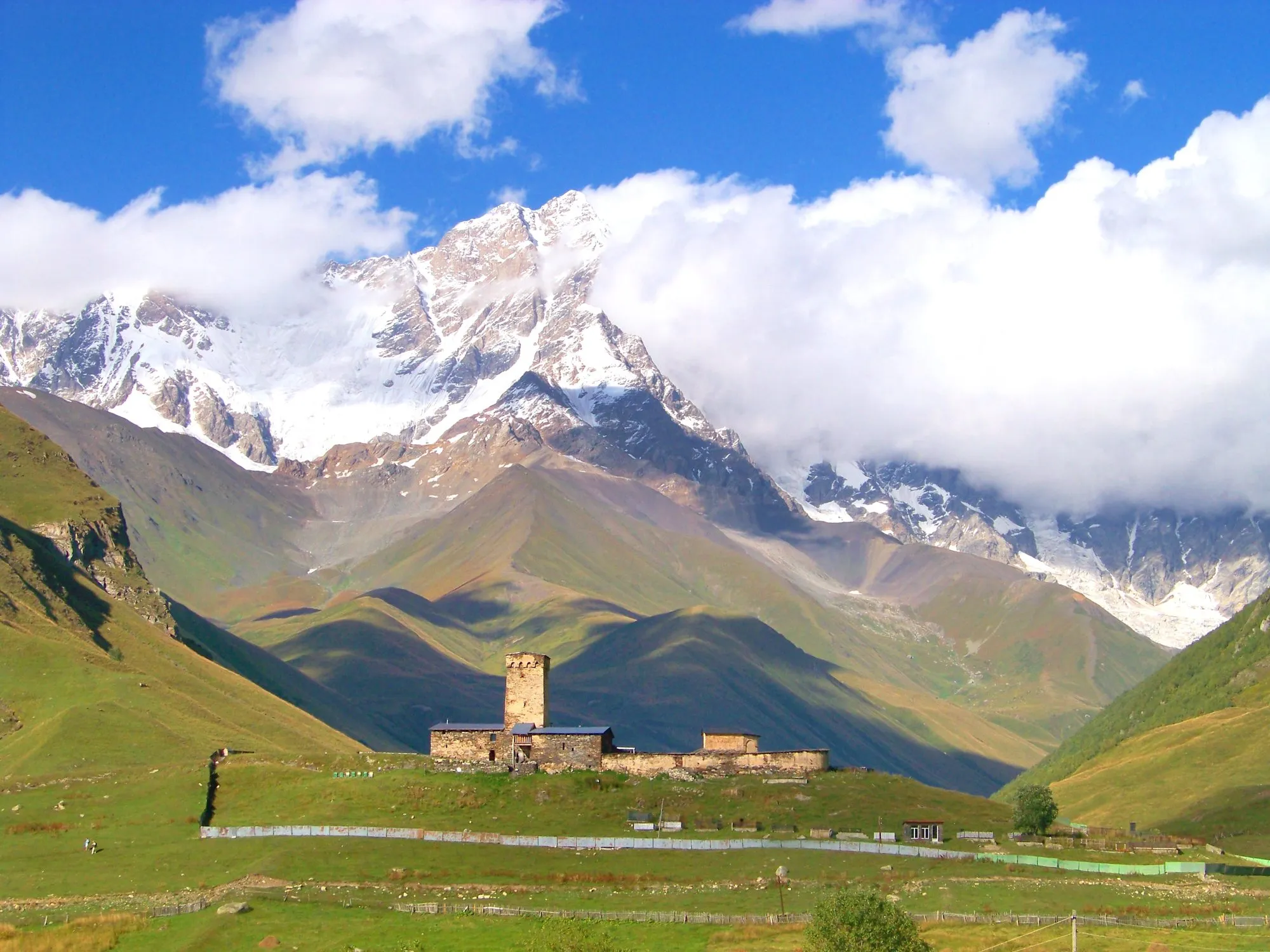 Lamaria church and its tower on a green hill below the snowy wall of Shkhara