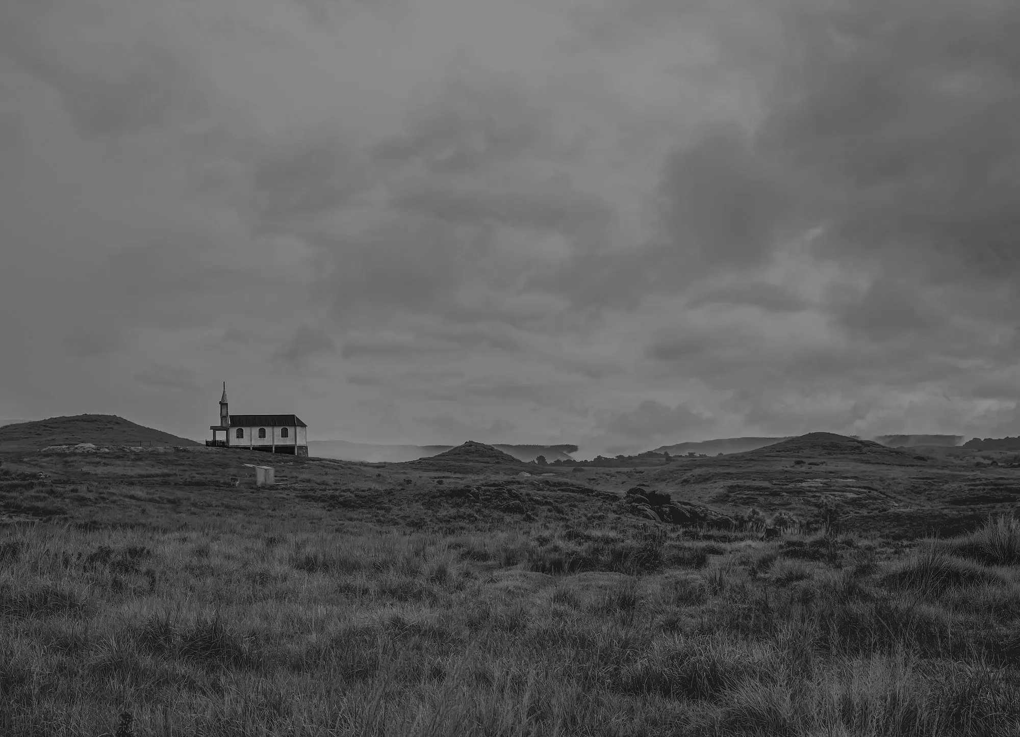 Black-and-white photo of a small church on grassland under heavy rain clouds at Mawsynram