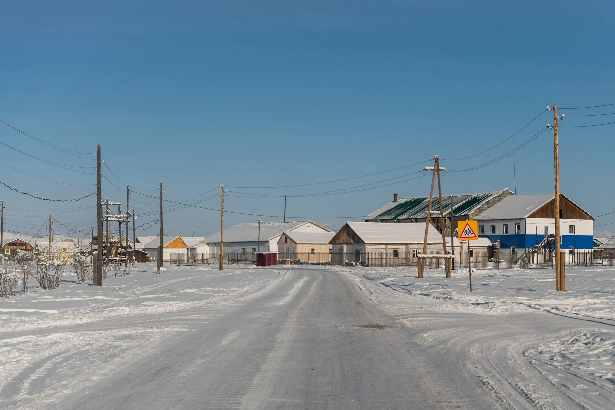 A snow-covered street with low wooden buildings and power lines in Oymyakon