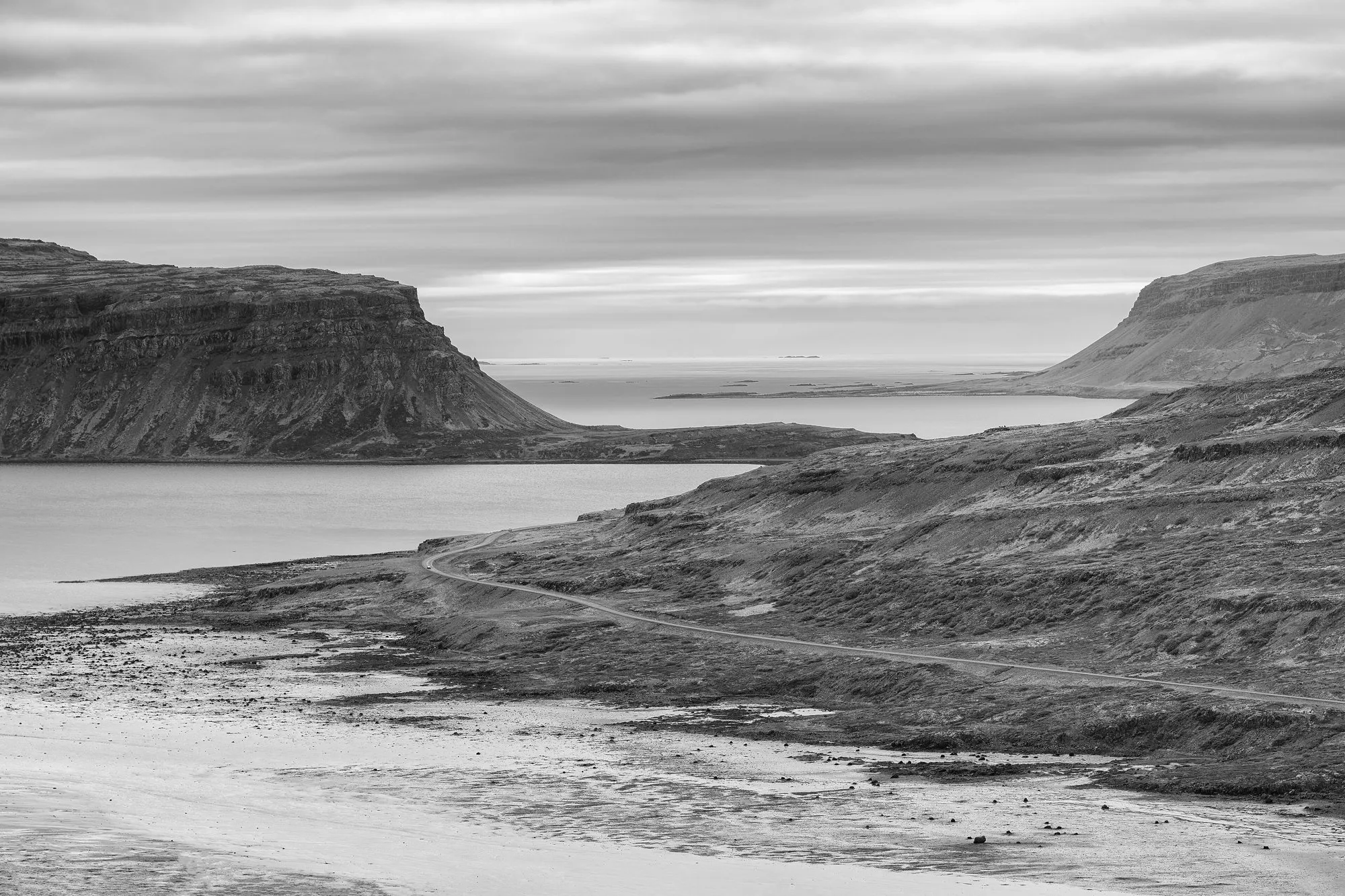 Black-and-white view of a road winding along a fjord between flat-topped mountains