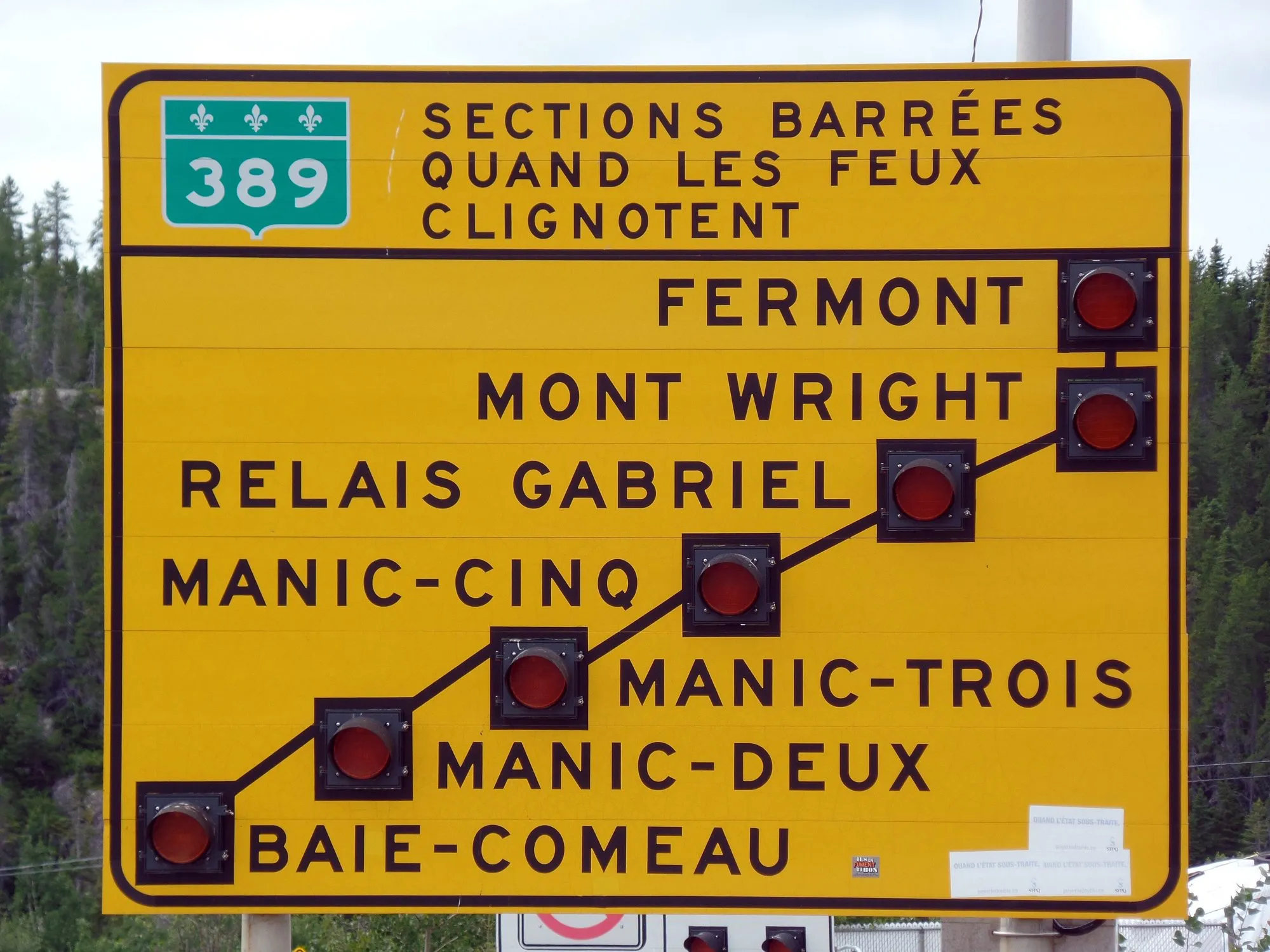 A yellow sign on Route 389 listing road sections that close when the lights flash