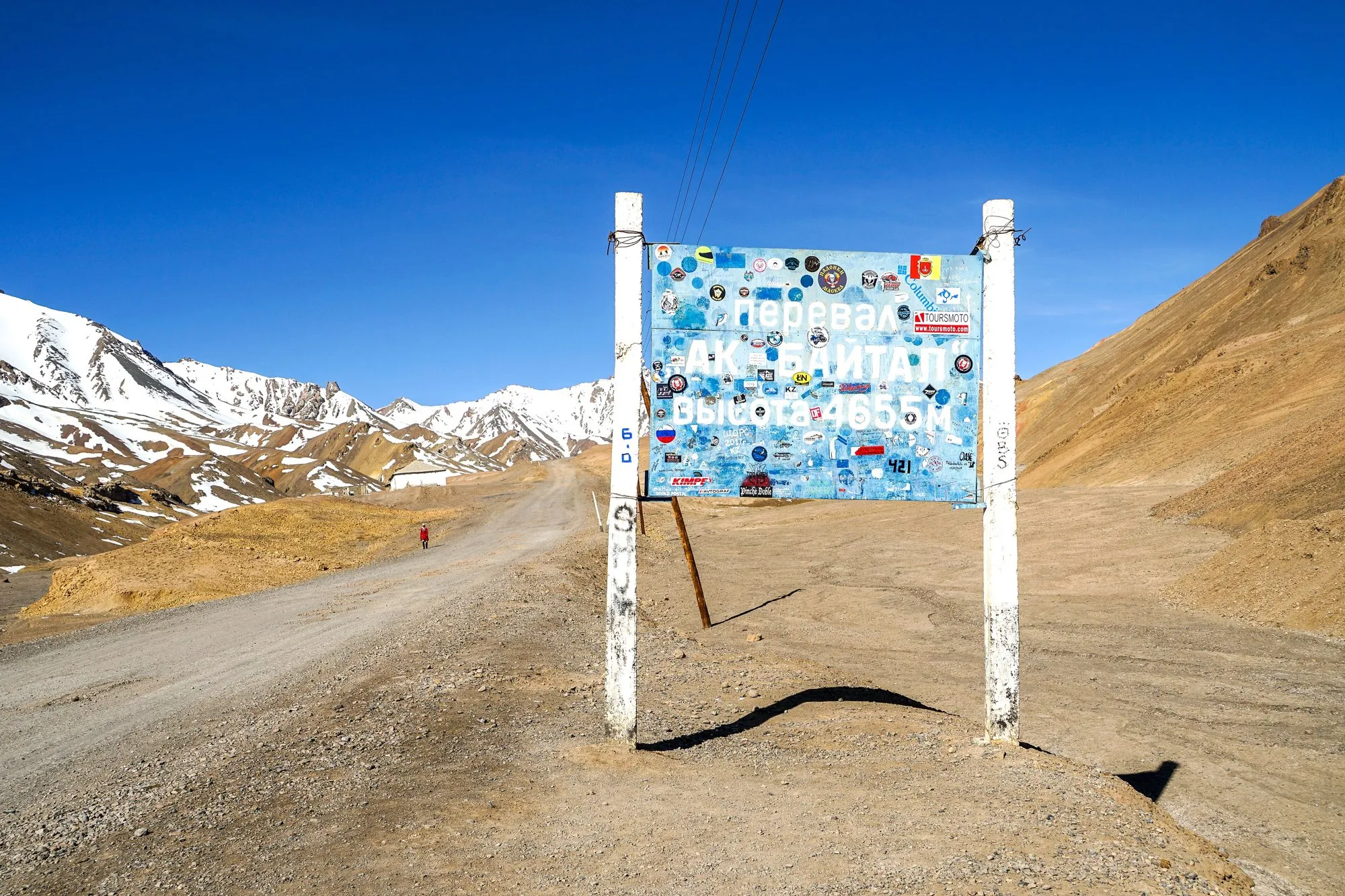 The sticker-covered Ak-Baital Pass sign beside the road with snowy mountains behind